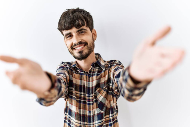 Homme hispanique avec la barbe debout sur fond isolé regardant la caméra souriant à bras ouverts pour étreindre. expression joyeuse embrassant le bonheur.  - Photo, image