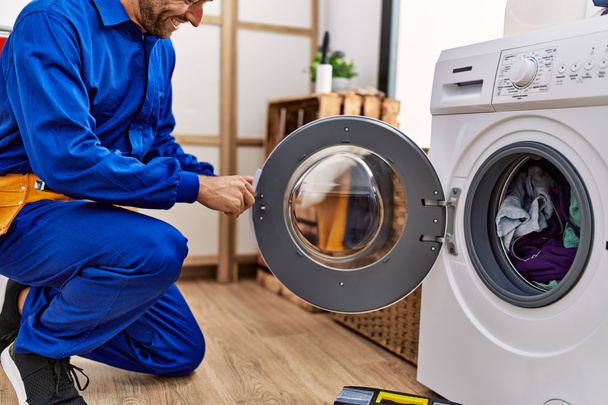 Young hispanic man wearing handyman uniform repairing washing machine at laundry room - Photo, Image