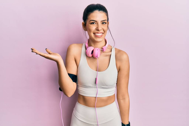 Jonge Latijns-Amerikaanse vrouw in gymkleren en met behulp van een koptelefoon glimlachend vrolijke presentatie en wijzen met palm van de hand kijken naar de camera.  - Foto, afbeelding