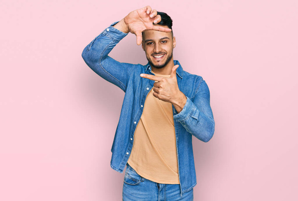 Junger arabischer Mann in lässiger Kleidung, der lächelnd mit Händen und Fingern ein fröhliches Gesicht macht. Kreativität und Fotografiekonzept.  - Foto, Bild