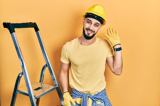 Handsome man with beard by construction stairs wearing hardhat waiving saying hello happy and smiling, friendly welcome gesture  - Φωτογραφία, εικόνα