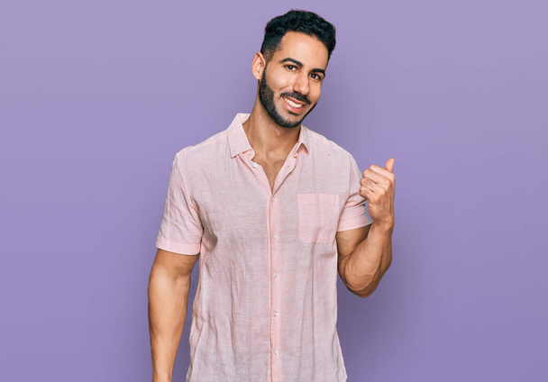 Hispanischer Mann mit Bart trägt lässiges Hemd, lächelt mit glücklichem Gesicht und zeigt mit erhobenem Daumen zur Seite.  - Foto, Bild