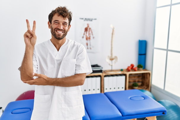 Молодой красивый физиотерапевт, работающий в клинике болеутоляющих, улыбаясь счастливым лицом, подмигивает в камеру, делая знак победы пальцами. Номер два.  - Фото, изображение