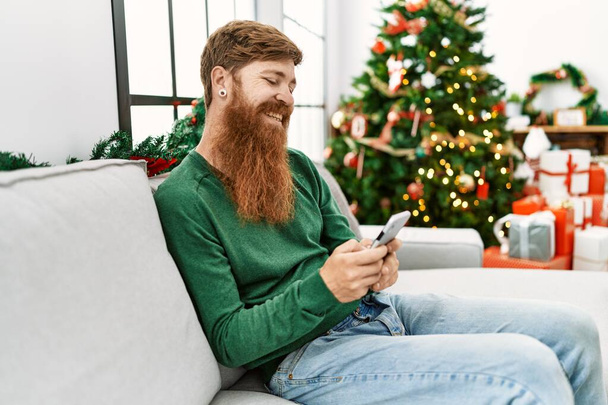 Νεαρός κοκκινομάλλης άντρας που χρησιμοποιεί smartphone κάθεται δίπλα στο χριστουγεννιάτικο δέντρο στο σπίτι - Φωτογραφία, εικόνα