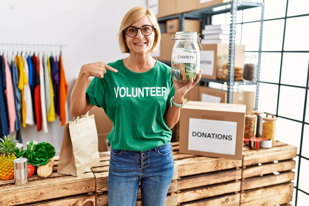 Blonde Frau mittleren Alters, die ein freiwilliges T-Shirt mit Spendengeldern trägt und mit dem Finger auf eine sich selbst lächelnde glücklich und stolz zeigt  - Foto, Bild