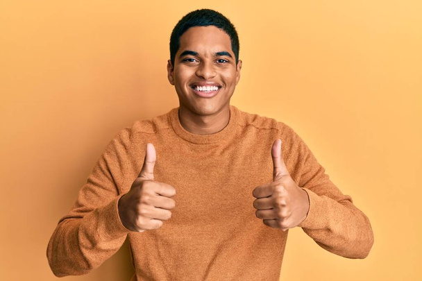 手で積極的なジェスチャーを行うカジュアルな冬のセーターの成功のサインを着て若いハンサムなヒスパニック系の男性は、親指を笑顔と幸せ。陽気な表情と勝者のジェスチャー.  - 写真・画像