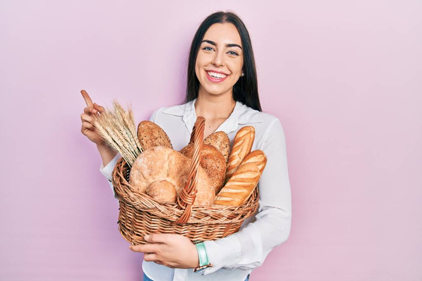 Beautiful woman with blue eyes holding wicker basket with bread smiling happy pointing with hand and finger to the side  - Foto, imagen