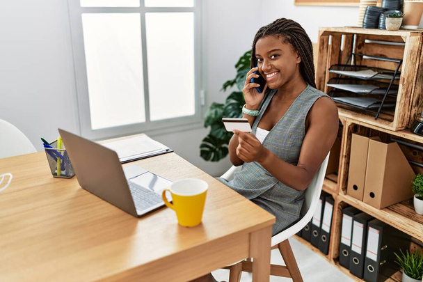 Trabajadora de negocios afroamericana joven hablando en el teléfono inteligente usando tarjeta de crédito en la oficina - Foto, imagen