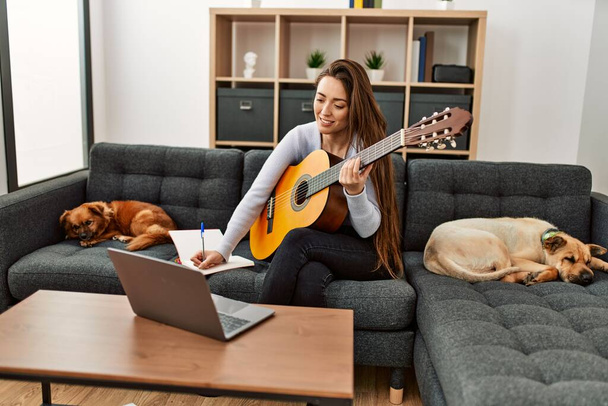 Nuori latino nainen ottaa online kitara luokka istuu sohvalla koirien kanssa kotona - Valokuva, kuva