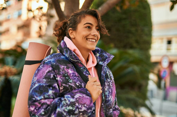 Młoda Latynoska sportowa kobieta uśmiechająca się szczęśliwa trzymając matę do jogi w parku. - Zdjęcie, obraz