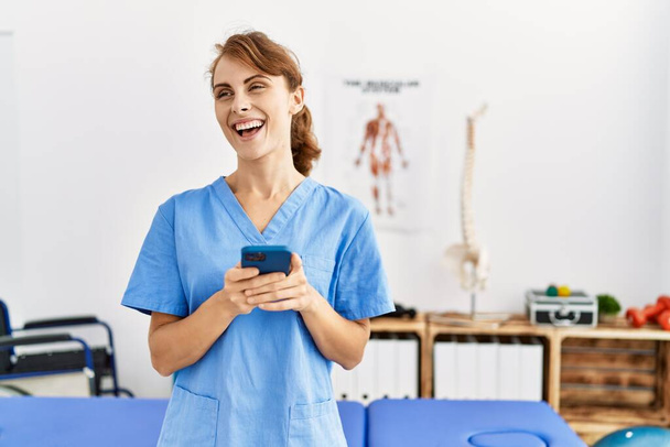 Junge kaukasische Physiotherapeutin lächelt glücklich mit Smartphone in der Klinik. - Foto, Bild