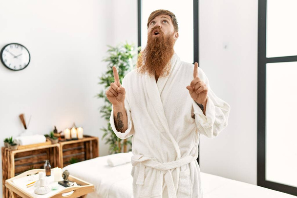 Redhead man with long beard wearing bathrobe at wellness spa amazed and surprised looking up and pointing with fingers and raised arms.  - Fotografie, Obrázek