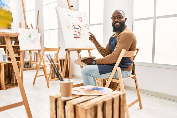 Afrikkalainen amerikkalainen taiteilija mies maalaus kankaalle art studio hymyillen onnellinen osoittaa käsin ja sormella  - Valokuva, kuva