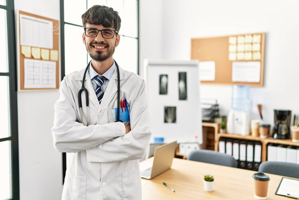 Nuori latino lääkäri mies hymyilee luottavainen kädet ristissä ele klinikalla - Valokuva, kuva