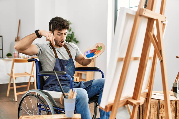 Sanat stüdyosunda tekerlekli sandalyede oturmuş reklamını gösteren parmaklarıyla işaret eden, şaşkın suratlı ve ağzı açık genç bir İspanyol.  - Fotoğraf, Görsel