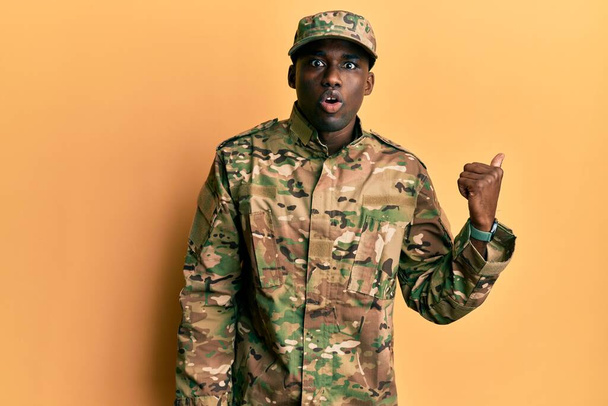 Young african american man wearing army uniform surprised pointing with hand finger to the side, open mouth amazed expression.  - Фото, изображение