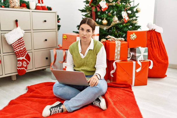 Junge schöne hispanische Frau sitzt mit Laptop am Weihnachtsbaum und sieht schläfrig und müde aus, erschöpft von Müdigkeit und Kater, faule Augen am Morgen.  - Foto, Bild