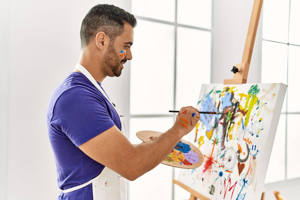Молодой латиноамериканец улыбается уверенно рисуя в художественной студии - Фото, изображение