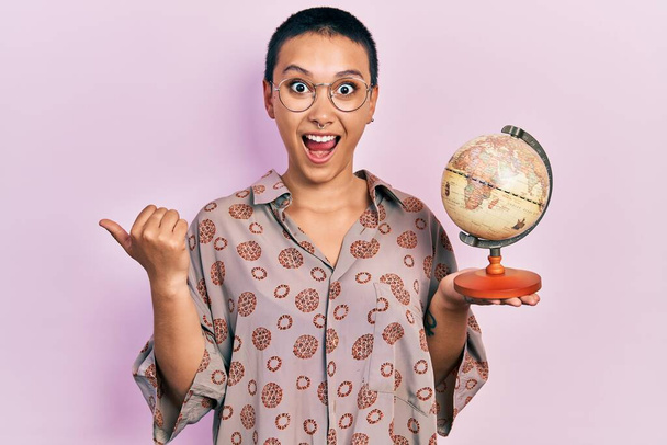 Όμορφη ισπανόφωνη γυναίκα με κοντά μαλλιά κρατώντας μικρή μπάλα του κόσμου δείχνοντας τον αντίχειρα μέχρι το πλάι χαμογελώντας ευτυχισμένη με ανοιχτό στόμα  - Φωτογραφία, εικόνα