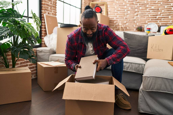 Young african american man unpacking books cardboard box at new home - Foto, imagen