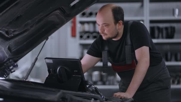 Мужчина-механик использует планшетный компьютер с программным обеспечением для диагностики дополненной реальности. Специалист осматривает автомобиль, чтобы найти сломанные компоненты внутри моторного отсека. Современный автосервис. - Кадры, видео