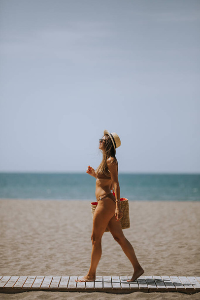 Красивая молодая женщина в бикини с соломенной сумкой на пляже в летний день - Фото, изображение
