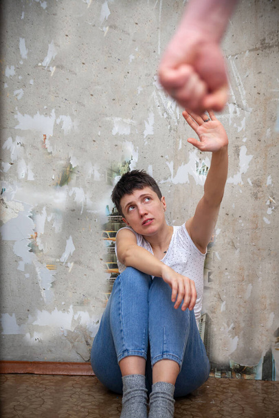 Eine verängstigte Frau sitzt auf dem Boden an der Wand und bedeckt ihr Gesicht mit den Händen eines aggressiven Mannes. Häusliche Gewalt. - Foto, Bild