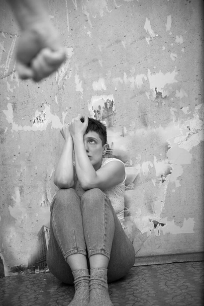 Испуганная женщина, сидящая на полу напротив стены, закрывая лицо руками от агрессивного мужчины. Бытовое насилие. - Фото, изображение