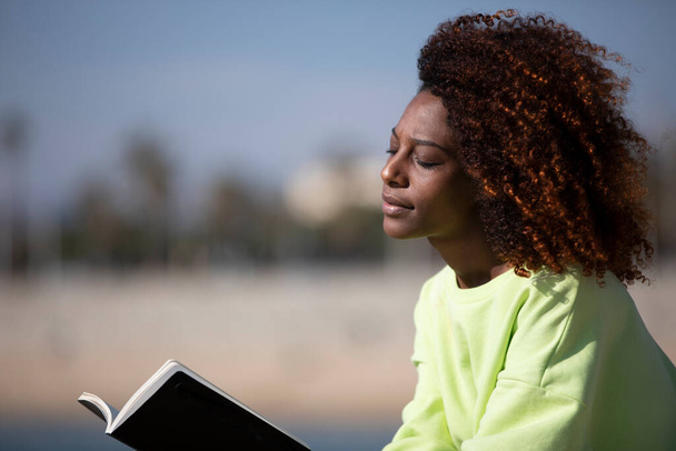 Young curly afro woman sitting on a breakwater holding a book while reading outdoors - Foto, afbeelding