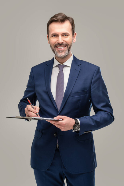 Happy handsome businessman holding clipboard and looking at camera, posing on grey studio background. Successful people. Insurance agent. Real estate broker - Фото, изображение