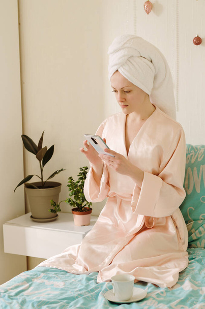 Jonge gelukkige vrouw met een handdoek op haar hoofd zittend in bed in een pyjama in een gezellig huis. Morgenkoffie drinken. Schoonheidsroutine. - Foto, afbeelding