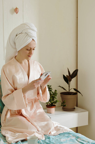 Jonge gelukkige vrouw met een handdoek op haar hoofd zittend in bed in een pyjama in een gezellig huis. Morgenkoffie drinken. Schoonheidsroutine. - Foto, afbeelding