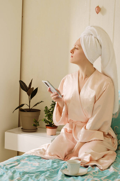 Young happy woman with a towel on her head sitting in bed in pajamas in a cozy home. Drinking morning coffee. Beauty routine. - 写真・画像