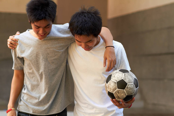 Хорошая игра. Два азиатских мальчика ходят, обнимая друг друга, держа футбольный мяч.. - Фото, изображение