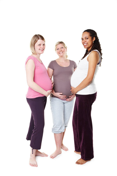 Ich fühle mich schon beschützt. Drei schwangere Frauen lächeln breit in die Kamera, während sie vor weißem Hintergrund stehen und den Bauch halten. - Foto, Bild