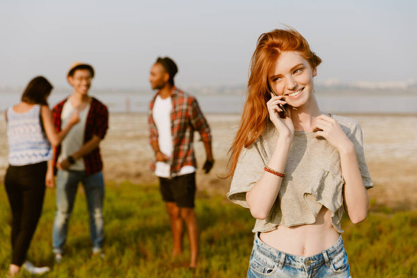 Young ginger woman smiling and talking on cellphone while resting with his friends outdoors - Photo, Image