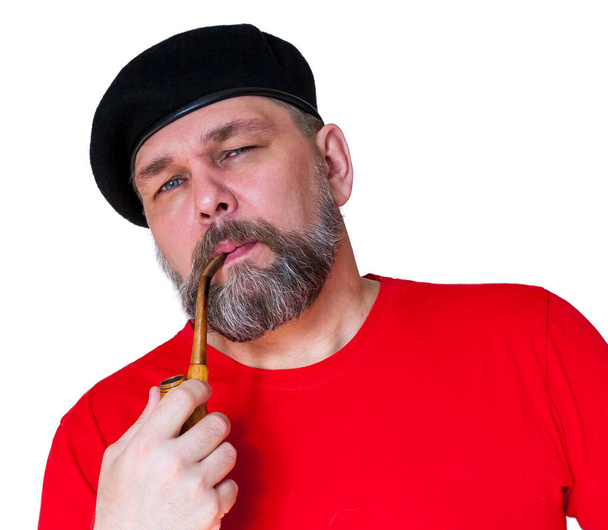 Een man van middelbare leeftijd met een grijze baard en een pijp. Baard man in rood t-shirt en zwarte baret, geïsoleerd op wit. Een rustige man van middelbare leeftijd rookt een pijp. - Foto, afbeelding