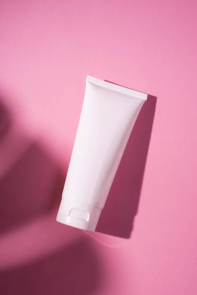 Plastic white tube for cream or lotion. Skin care or sunscreen cosmetic in top view on pink background with palm leaves shadow. Beauty concept for face care - Fotó, kép