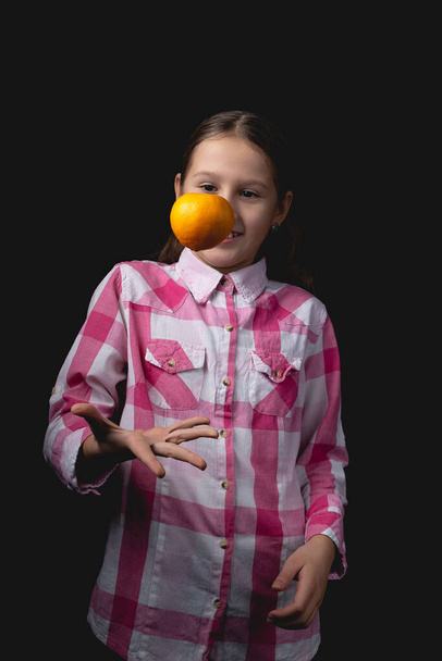 A kis aranyos lány mandarinnal pózol a stúdióban. Fénykép elszigetelt fekete háttérrel egy fényforrás. A gyerek hülyéskedik azzal, hogy egy mandarint dob a levegőbe.. - Fotó, kép