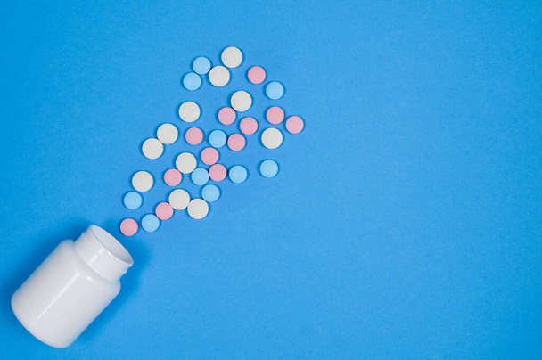 Weiße, rosa und blaue Pillen aus einer weißen Flasche auf blauem Hintergrund. - Foto, Bild