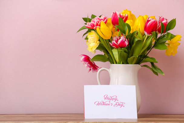 Caraffa con bellissimi tulipani e biglietto di auguri con testo GIORNO DELLA FELICE DONNA sul bancone della cucina vicino alla parete colorata - Foto, immagini