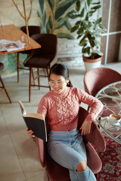 Una chica asiática relajada se sienta en una cafetería y lee un libro. - Foto, imagen