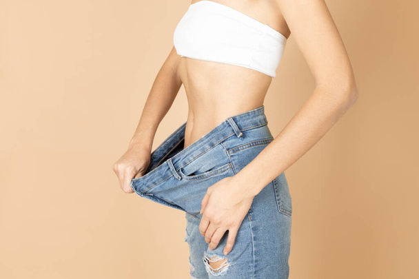 Fit woman having ripped jeans and white bra on stretching jeans to show belly after weight loss. Keeping diet and exercising. Aiming for improving health and getting fit - Foto, afbeelding
