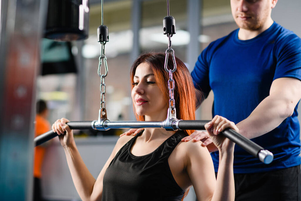 Attrayant femme entraînement d'exercice muscles des mains dans l'intérieur de la salle de gym moderne avec entraîneur masculin et machine avec chaînes - Photo, image