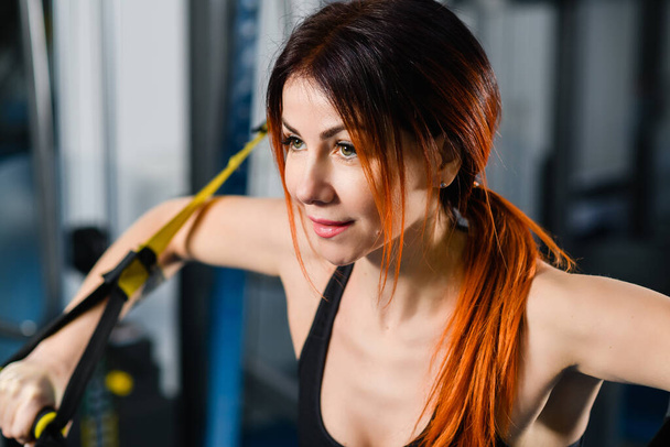 Young athletic woman with perfect body in sportswear doing push ups with trx fitness straps in the gym - Fotografie, Obrázek