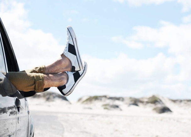 Taking a break to stretch my legs. Cropped image of two feet wearing sneakers sticking out of a parked car window with dunes in the background. - 写真・画像