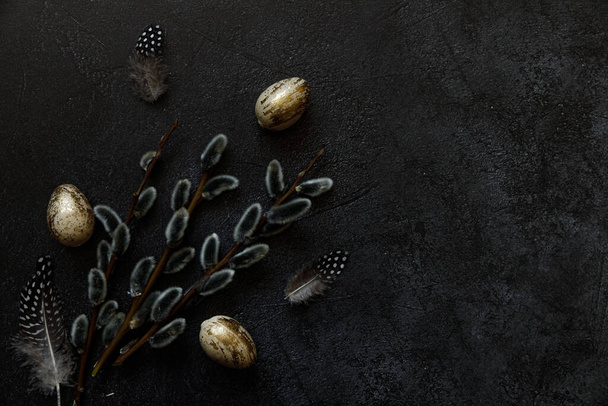 Happy Easter concept. Preparation for holiday. Golden decorated easter eggs willow feather on trendy grunge scratched dark black shale background. Simple minimalism flat lay top view, copy space - 写真・画像