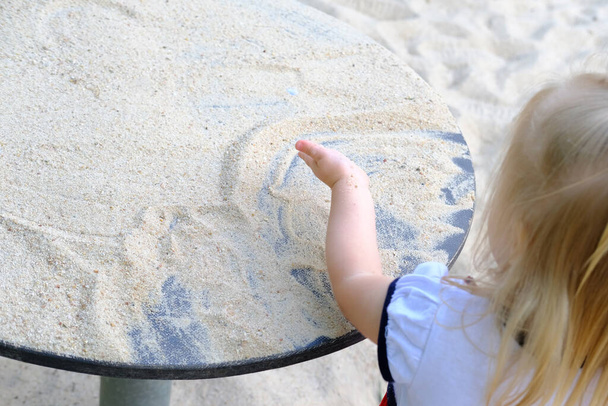 small child, 2-year-old girl plays in summer with sand on seashore, in sandbox, on playground, concept of development of fine motor skills, build sand castles, summer vacation, fun on beach - Photo, image