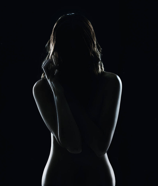 Sensueel silhouet. Silhouet van een naakte jonge vrouw tegen een donkere bakgrond. - Foto, afbeelding