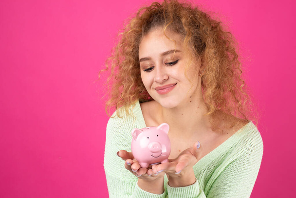 A cute young girl with curly red hair holds a piggy bank, a pink piglet in her hands. The concept of wealth and safety of money. - Fotografie, Obrázek